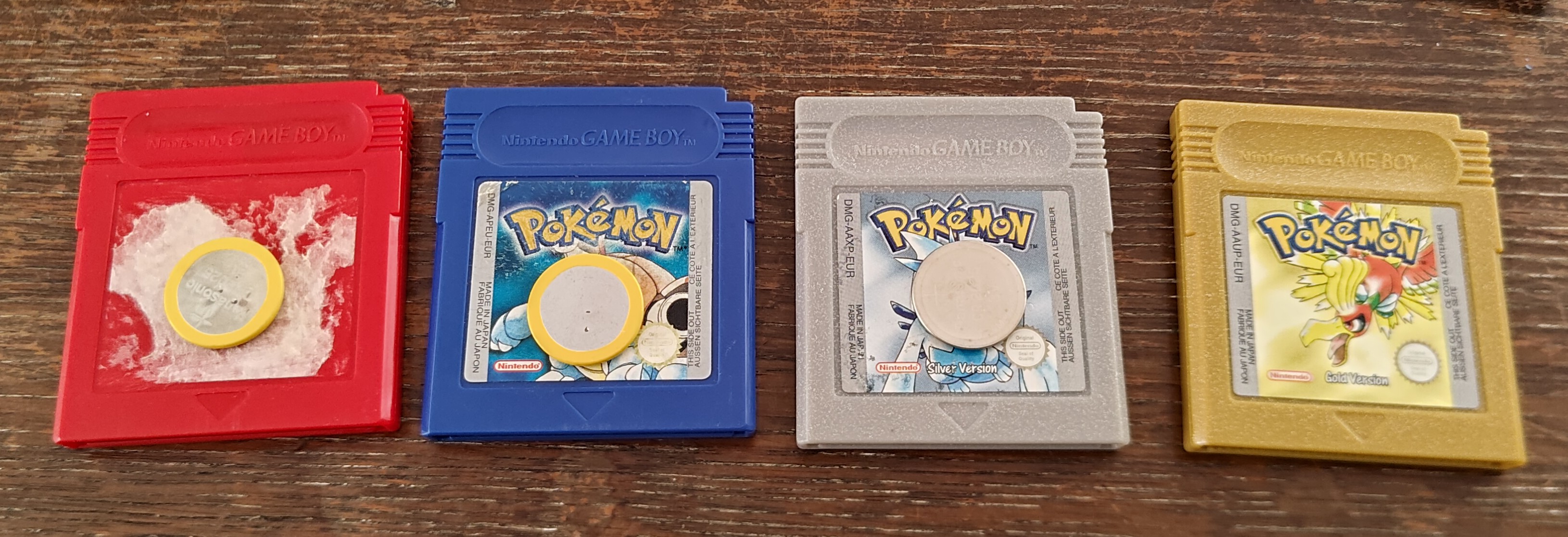 Cartridges for Pokémon Red, Blue, Silver, and Gold. The first three have the recently-removed batteries on top of their labels (or lack-of-label for Red). The batteries for Red and Blue both have the original yellow edge.
